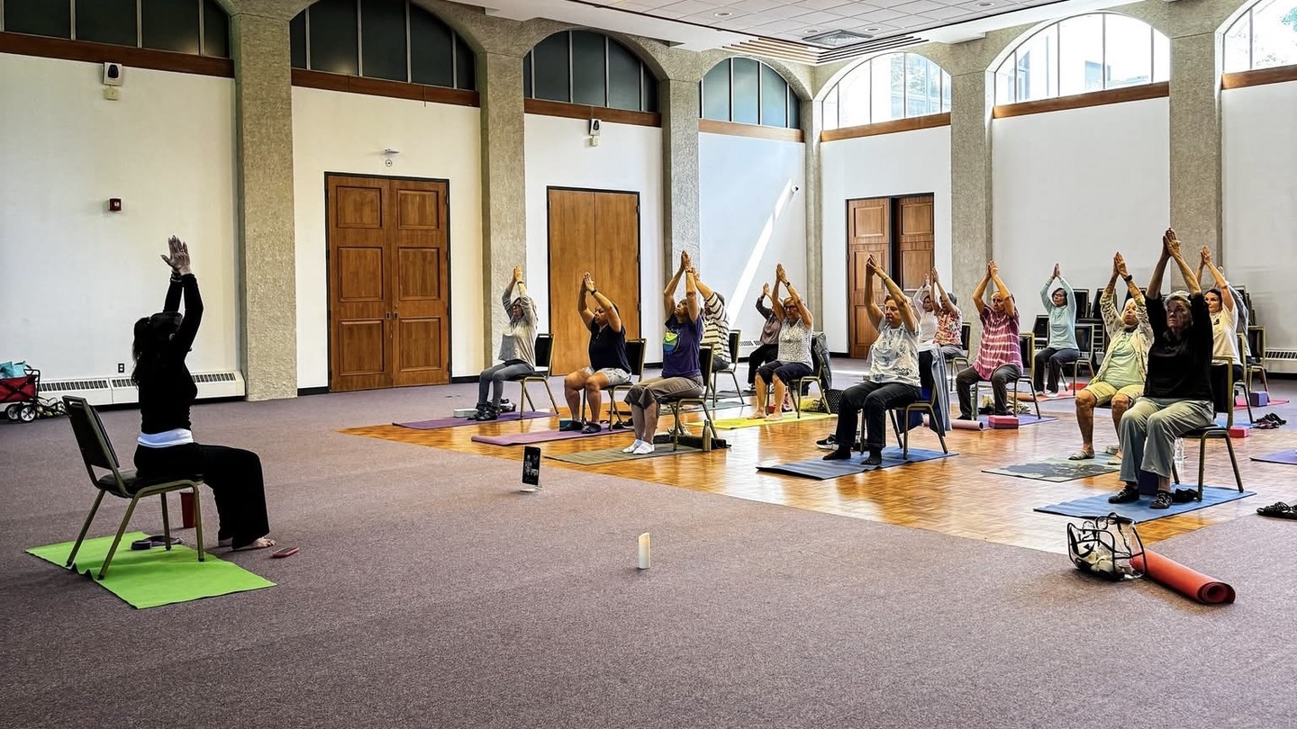Miss Susan leading a chair yoga class with adults seated in a community room.